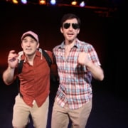 Stage production photo featuring two performers running toward the audience on a theater stage. One performer wears sunglasses and a plaid shirt, while the other wears a pink cap, backpack, and short-sleeve shirt. Colored stage lights illuminate the dark background.