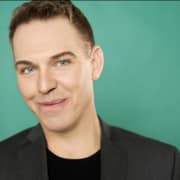 Professional headshot of a person with short hair wearing a dark blazer over a black shirt. The portrait is framed from the shoulders up against a teal background with soft studio lighting.