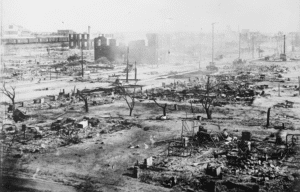 Black-and-white photograph of widespread destruction in Tulsa, Oklahoma, following the 1921 Tulsa Race Massacre. Burned foundations, scattered debris, charred trees, and the ruins of buildings stretch across the landscape, with damaged structures and streets visible in the background.