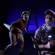 Stage production photo featuring two performers in casual outdoor-themed costumes on a dark stage lit with blue and purple lighting. One performer sits in the foreground holding a flashlight, while another stands behind wearing a bicycle helmet. A ladder and oversized prop are visible in the background, creating a parody-adventure atmosphere.
