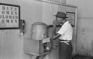 Black-and-white photograph showing a person drinking from a water fountain labeled “Colored” beneath signs reading “White Women” and “Colored Women,” illustrating racial segregation in a public facility during the Jim Crow era. A large mirror and paneled walls are visible in the background.