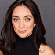 Professional headshot of a person with long dark hair wearing a black sleeveless turtleneck top. The portrait is framed from the shoulders up against a dark gray background, with the subject facing the camera under soft studio lighting.