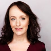 Professional headshot of a person with shoulder-length dark hair wearing a burgundy top. The portrait is set against a light gray background and framed from the shoulders up, with even studio lighting and a clear focus on the face.