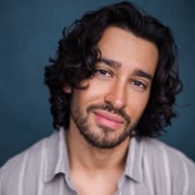 Professional headshot of a person with shoulder-length curly hair and a short beard, wearing a light-colored button-front shirt. The portrait is tightly framed against a blue studio background, with soft lighting and direct eye contact with the camera.