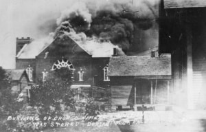 Black-and-white photograph of a church engulfed in flames during the 1921 Tulsa Race Massacre. Thick smoke rises from the roof as nearby buildings stand in the foreground. Handwritten text on the image identifies the church and notes that ammunition was stored there during the violence.