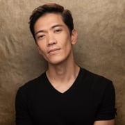 Professional headshot of a person wearing a black V-neck shirt, photographed against a textured tan background. The image is cropped from the chest up, with the subject facing the camera and slightly tilting their head. Soft studio lighting highlights the face and creates subtle shadows against the backdrop.
