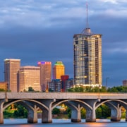 Downtown Tulsa, Oklahoma skyline at dusk, featuring a tall cylindrical high-rise tower and illuminated office buildings behind a multi-arched concrete bridge spanning the Arkansas River under dramatic blue-gray storm clouds.