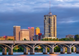 Downtown Tulsa, Oklahoma skyline at dusk, featuring a tall cylindrical high-rise tower and illuminated office buildings behind a multi-arched concrete bridge spanning the Arkansas River under dramatic blue-gray storm clouds.