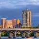 Downtown Tulsa, Oklahoma skyline at dusk, featuring a tall cylindrical high-rise tower and illuminated office buildings behind a multi-arched concrete bridge spanning the Arkansas River under dramatic blue-gray storm clouds.