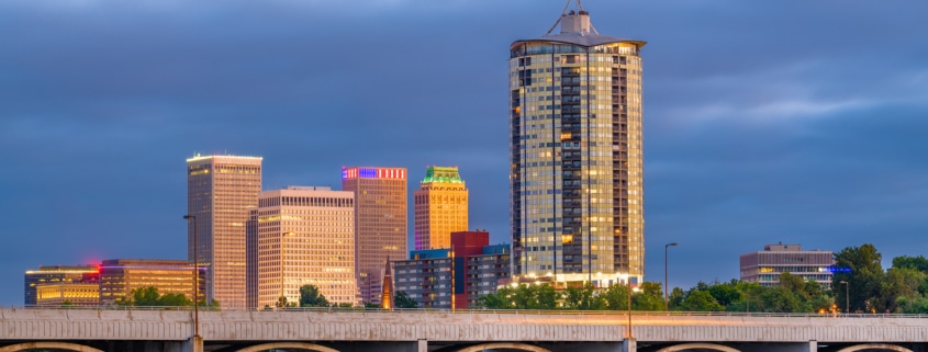 Downtown Tulsa, Oklahoma skyline at dusk, featuring a tall cylindrical high-rise tower and illuminated office buildings behind a multi-arched concrete bridge spanning the Arkansas River under dramatic blue-gray storm clouds.