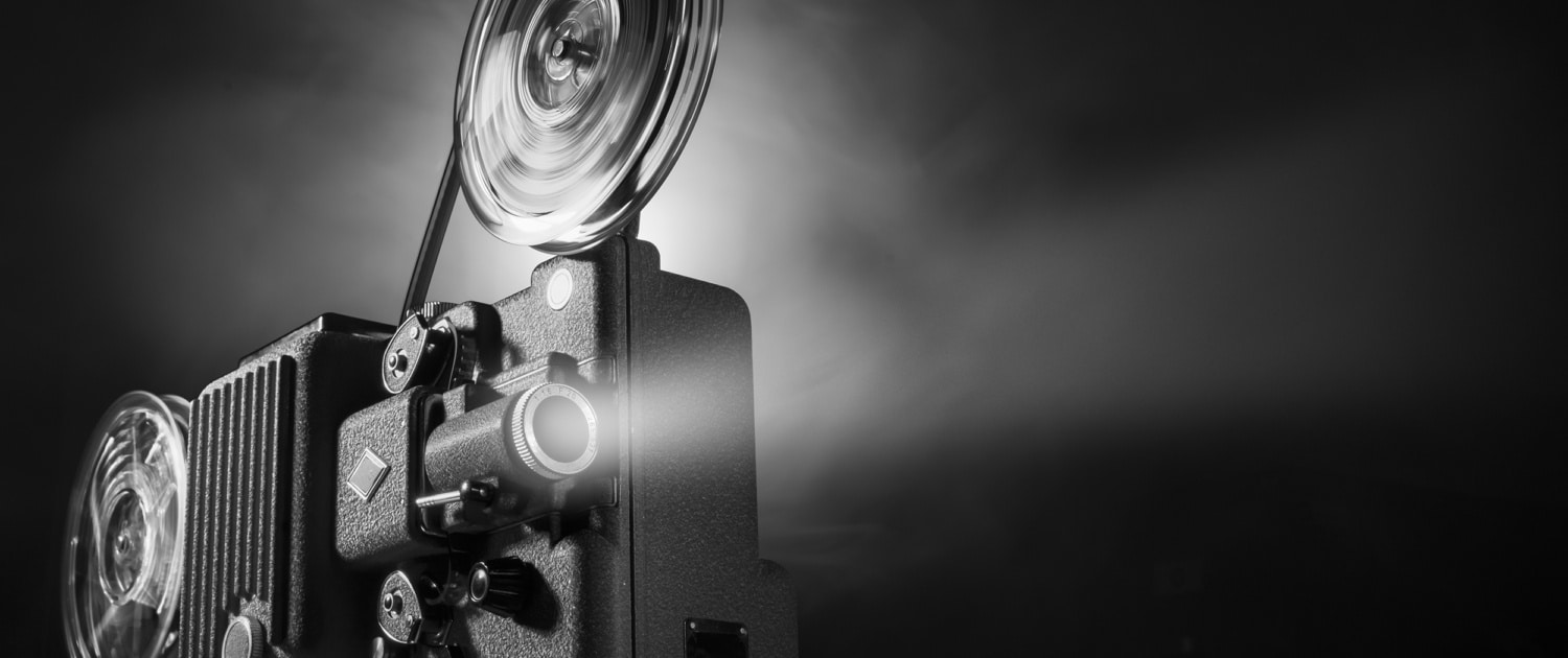 Black-and-white image of a vintage film projector with large spinning reels casting a bright beam of light into a dark space. The projector occupies the left side of the frame, while the illuminated projection beam extends across the image against a black background, emphasizing the equipment's mechanical details and cinematic atmosphere.