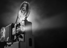 Black-and-white image of a vintage film projector with large spinning reels casting a bright beam of light into a dark space. The projector occupies the left side of the frame, while the illuminated projection beam extends across the image against a black background, emphasizing the equipment's mechanical details and cinematic atmosphere.