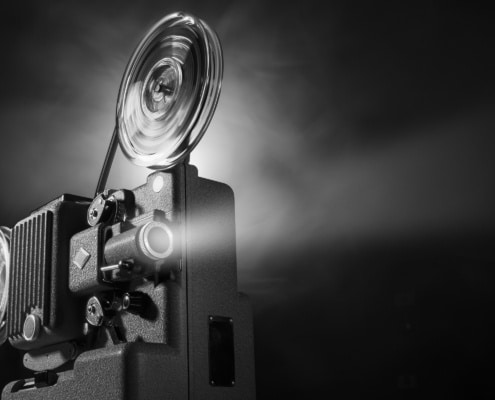 Black-and-white image of a vintage film projector with large spinning reels casting a bright beam of light into a dark space. The projector occupies the left side of the frame, while the illuminated projection beam extends across the image against a black background, emphasizing the equipment's mechanical details and cinematic atmosphere.