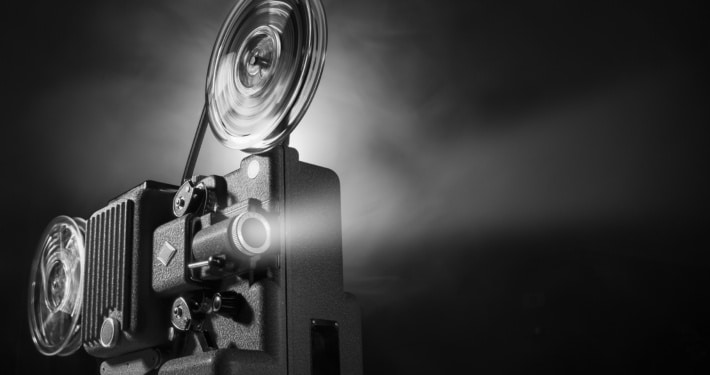 Black-and-white image of a vintage film projector with large spinning reels casting a bright beam of light into a dark space. The projector occupies the left side of the frame, while the illuminated projection beam extends across the image against a black background, emphasizing the equipment's mechanical details and cinematic atmosphere.