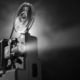 Black-and-white image of a vintage film projector with large spinning reels casting a bright beam of light into a dark space. The projector occupies the left side of the frame, while the illuminated projection beam extends across the image against a black background, emphasizing the equipment's mechanical details and cinematic atmosphere.
