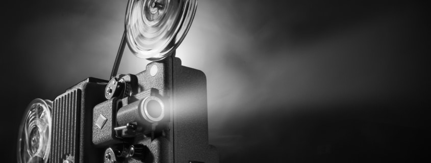 Black-and-white image of a vintage film projector with large spinning reels casting a bright beam of light into a dark space. The projector occupies the left side of the frame, while the illuminated projection beam extends across the image against a black background, emphasizing the equipment's mechanical details and cinematic atmosphere.