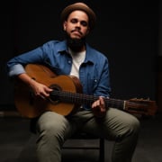 Musician seated with an acoustic guitar, wearing a denim shirt, light-colored pants, and a brown hat. The portrait is photographed against a dark background with studio lighting highlighting the instrument and performer.