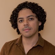 Professional headshot of a person with short curly hair wearing a brown button-front shirt. The portrait is framed from the shoulders up against a warm beige background with soft studio lighting.