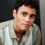 Professional headshot of a person with short curly hair wearing a light-colored collared shirt with vertical striped accents. The portrait is framed from the shoulders up against a dark neutral background with soft studio lighting.