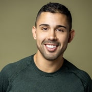 Professional headshot of a person with short dark hair wearing a dark green textured sweater. The portrait is framed from the shoulders up against an olive-green background with soft studio lighting.