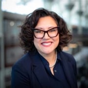 Professional headshot of a person with short curly hair wearing glasses, a dark blazer, and a pearl necklace. The portrait is framed from the shoulders up against a softly blurred office background with natural lighting.