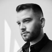 Black-and-white professional portrait of a person with short hair and a trimmed beard wearing a dark collared shirt. The image is framed from the shoulders up against a light background, with side lighting creating contrast and depth.