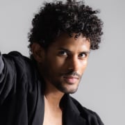 rofessional headshot of a person with short curly hair wearing a dark jacket. The portrait is closely cropped and photographed against a light gray background with soft studio lighting.