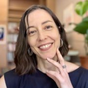 Professional headshot of a person with shoulder-length dark hair wearing a sleeveless navy top and silver earrings. The portrait is photographed indoors against a softly blurred office-like background with warm natural light.