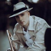 Portrait of a musician wearing a light-colored fedora and striped jacket while holding a trombone. The image is photographed outdoors with a softly blurred background and warm, natural lighting. Provide your feedback on BizChat