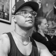 Black-and-white portrait of a person wearing a baseball cap, glasses, and a sleeveless shirt. The image is photographed indoors with framed artwork and softly blurred figures in the background.