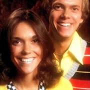 Vintage studio portrait of two people posing against a dark background. The person in front wears a bright yellow sweater with a black-and-white checkered collar and shoulder-length dark hair. The person behind wears a yellow collared shirt under a striped sweater with red, black, gray, and yellow bands. Both face the camera in a warmly lit 1970s-style promotional photograph.