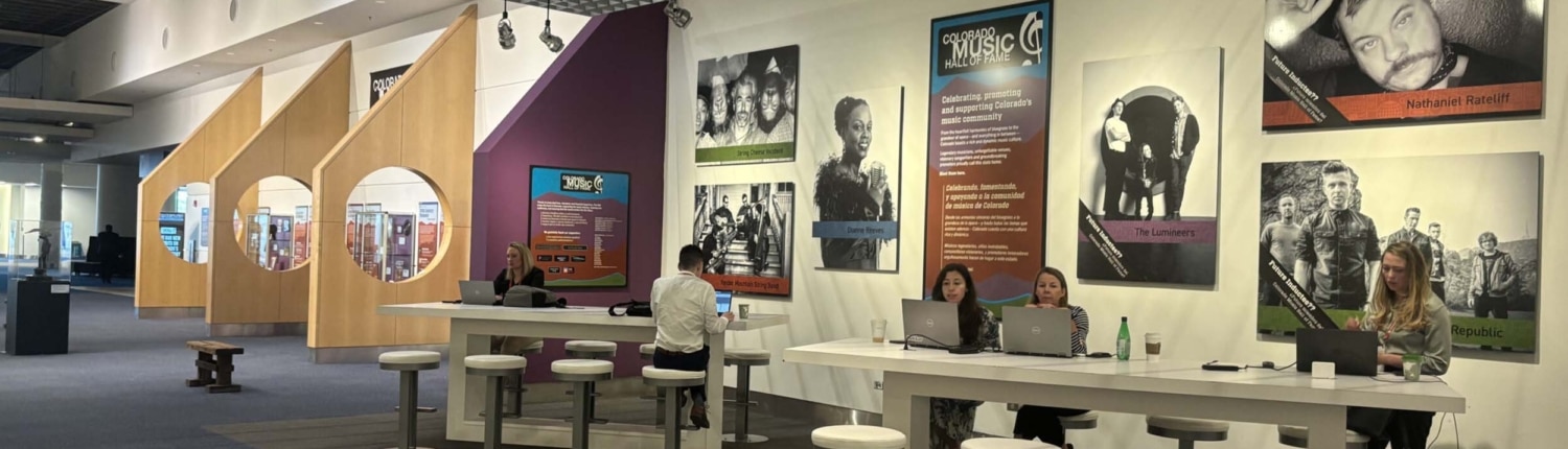 Interior of the Colorado Music Hall of Fame exhibit space, featuring large musician portraits on the wall, interactive worktables with stools and laptops, and a series of wooden display structures extending down a spacious gallery.