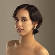 Professional headshot of a person with short dark hair wearing hoop earrings. The portrait is framed from the shoulders up against a warm beige background with soft studio lighting.