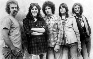 Black-and-white promotional portrait of five members of a rock band standing side by side against a textured wall, wearing casual 1970s-era clothing and looking toward the camera.