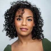 Professional headshot of a person with short curly hair wearing a green top and a delicate necklace. The portrait is framed from the shoulders up against a light gray background with soft, even lighting.
