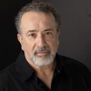 Professional headshot of a person with short gray hair and a gray beard wearing a black collared shirt. The portrait is framed from the shoulders up against a dark neutral background with studio lighting.