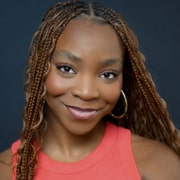Professional headshot of a person with long braided hair, hoop earrings, and a sleeveless coral top. The portrait is framed from the shoulders up against a dark blue background with even studio lighting.