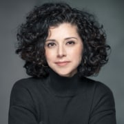 Professional headshot of a person with shoulder-length curly dark hair wearing a black turtleneck sweater. The portrait is framed from the shoulders up against a gray studio background with soft, even lighting.