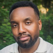 Professional outdoor headshot of a person with short hair and a neatly trimmed beard wearing a light-colored collared shirt. The portrait is framed from the shoulders up with a softly blurred green background and natural lighting.