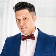 Professional headshot of a person wearing a blue suit jacket, white shirt, and burgundy bow tie. The portrait is framed from the chest up against a bright, softly lit background.