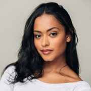 Professional headshot of a person with long dark hair wearing a light-colored top. The portrait is framed from the shoulders up against a light gray background with soft, even lighting.