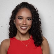 Professional headshot of a person with long curly hair wearing a sleeveless red top and a delicate necklace. The portrait is framed from the chest up against a light textured background with soft, even lighting.