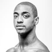 Black-and-white studio portrait of a person with a closely cropped hairstyle. The image is framed from the shoulders up against a light gray background with high-contrast lighting and a side-facing pose.