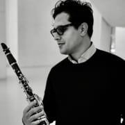 Black-and-white portrait of a musician holding a clarinet. The person wears glasses and a dark sweater over a light collared shirt, photographed in profile against a minimalist architectural background.