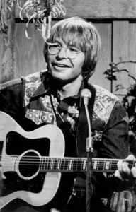 Black-and-white photograph of a musician seated with an acoustic guitar and singing into a microphone during a performance, wearing round glasses and a patterned shirt.