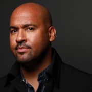 Professional headshot of a bald person with a goatee wearing a dark jacket over a collared shirt. The portrait is framed from the shoulders up against a dark gray background with dramatic studio lighting.