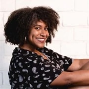 Professional portrait of a person with curly hair wearing a patterned black top. The subject is seated against a white brick wall, with the image framed from the waist up.