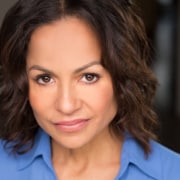 Professional headshot of a person with shoulder-length wavy hair wearing a blue collared shirt. The portrait is closely cropped against a softly blurred dark background with natural-looking lighting.
