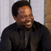 Professional portrait of a person wearing a black shirt and vest, seated indoors against a white brick wall. The image is framed from the waist up with warm natural lighting and a softly blurred background.