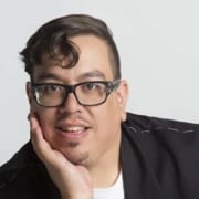 Professional headshot of a person wearing glasses and a dark jacket, resting their face on one hand. The portrait is closely cropped against a light gray background with soft, even lighting.